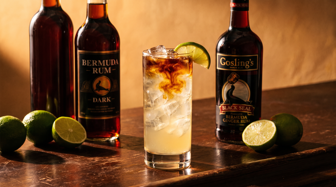 A classic Dark 'n' Stormy rum highball in a tall glass over ice with dark rum floating through pale ginger beer and a fresh lime wedge, beside dark Bermuda rum bottles and limes on a dark wood bar
