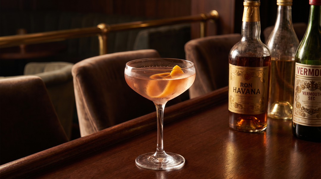 A classic El Presidente cocktail in a chilled coupe glass with an orange peel twist, beside white and gold Cuban-style rum bottles and dry vermouth on a dark-wood bar