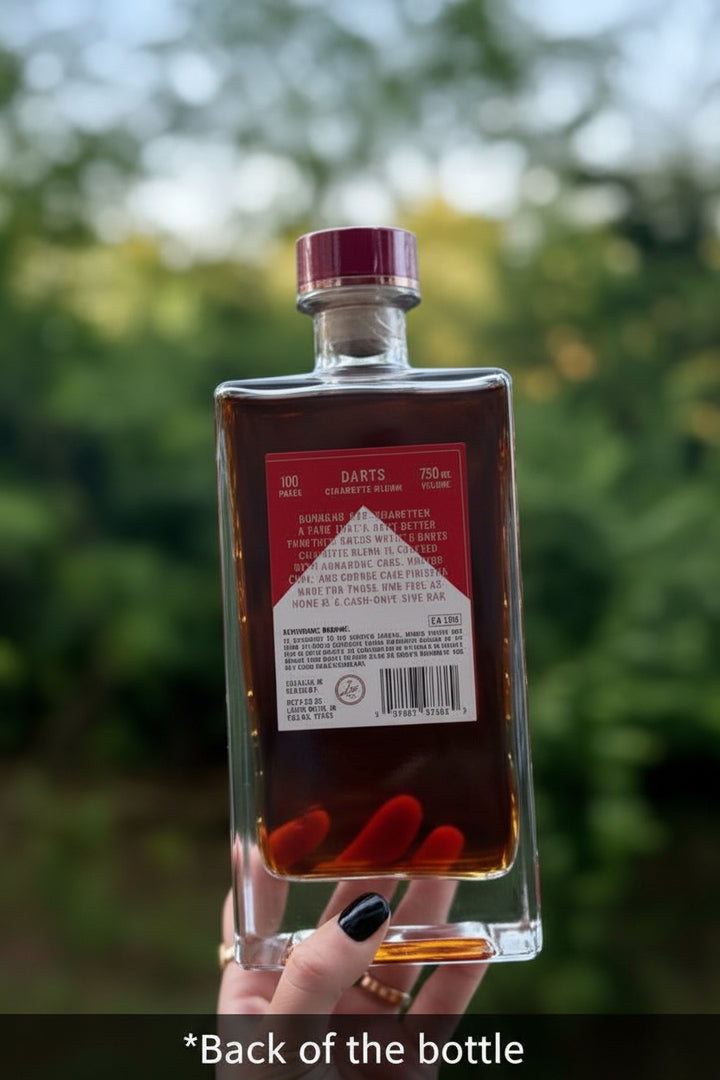 A hand holds a square glass bottle of Darts Whiskey against a blurred green outdoor background. The bottle contains dark amber liquid and has a red cap, with the back label showing product details.